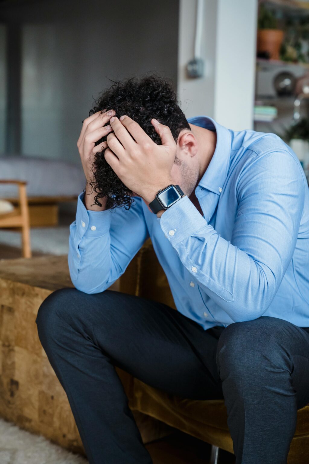 A person showing signs of stress, sitting with hands on head indoors wearing a blue shirt.